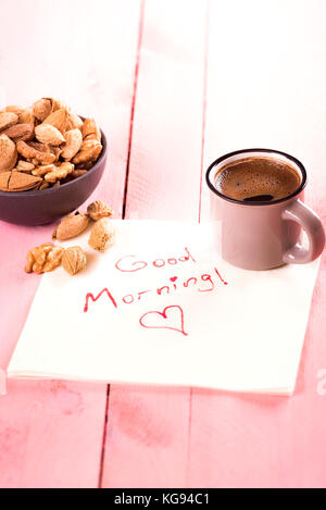 Frühstück mit einer Schale, in der Schale Mandeln, Walnüsse und einer aromatischen Tasse Kaffee auf einer Serviette mit den Worten "Guten Morgen geschrieben, auf einer Rosa woode Stockfoto