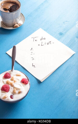 Weiß Serviette mit einem Entwurf für eine Aufgabenliste, indem eine Schüssel mit Joghurt mit Müsli und Himbeere und einer Tasse aromatischen Kaffee umgeben, auf einem blauen Hintergrund Stockfoto