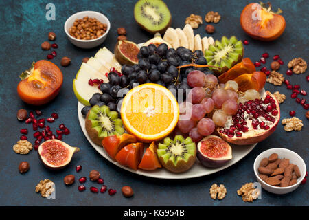 Platte mit frischem Obst und Nüsse: Orangen, Trauben, Kiwi, Kaki, Feigen, Mandeln, Walnüsse, Pinienkerne auf einem blauen Hintergrund Stockfoto