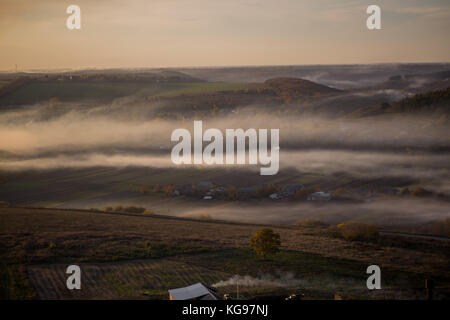 Eine Luftaufnahme von einem Dorf zwischen Feldern und Wäldern, auf dem am Abend Nebel verbreitet entfernt Stockfoto