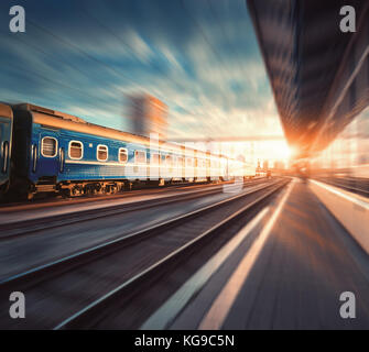 Schönen Zug mit blauen Wagen in Bewegung am Bahnhof bei Sonnenuntergang. Gewerbliche Anzeigen mit moderner Zug, Bahn, Bahnsteig, Gebäude, Stockfoto