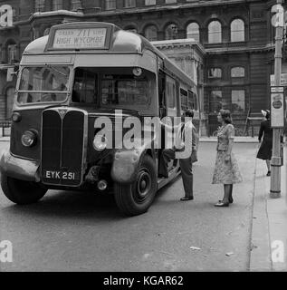 1950s, historisches Bild mit einem ausländischen Gentleman und einer Dame hinter ihm in Charing Cross, Central London, der darauf wartet, den 711 Green Line Bus über Oxford Circus in die Grafschaft High Wycombe in Buckinghamshire, England, Großbritannien, zu besteigen. Stockfoto