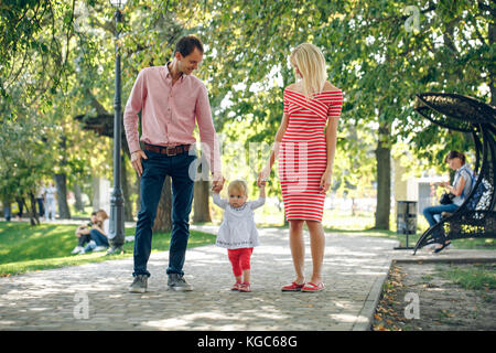 Familie Wandern im Park. Mama und Papa gehen Hand in Hand mit einem Baby. Eltern werden unterrichtet, Baby zu gehen Stockfoto