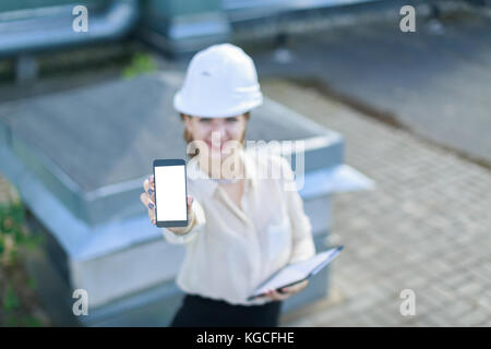 Attraktive businesslady in weiße Bluse, Watch, Helm und schwarzen Rock stand auf dem Dach und leeren Smartphone anzeigen Stockfoto