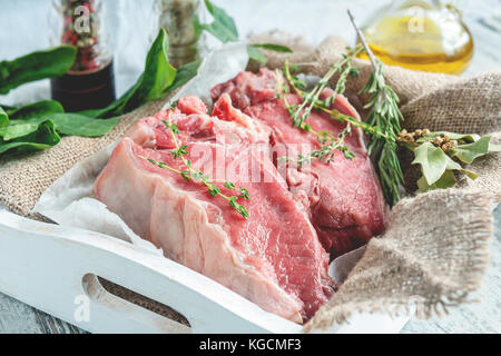 Teilstücke von Rindfleisch für das Grillen auf einem hölzernen Schneidebrett mit Spinat, Rosmarin und Kräutern der Provence für die Marinade in einer rustikalen Stil. Die horizontale Fram Stockfoto