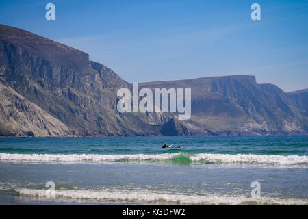 Surfer im Wasser auf der Suche nach Wellen mit spektakulären Minaun Cliffs im Hintergrund an einem Sandstrand von Keel Beach, Achill Island, County Mayo, Irland Stockfoto