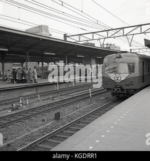 1950, historische, eine S-Bahn kommt auf einer Plattform, während ein anderer sehen wir japanische Pendler eine geordnete Warteschlange auf ihre Bahn anreisen, Tokio, Japan gebildet haben. Stockfoto