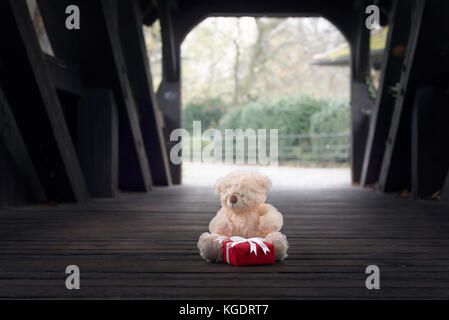 Süße gefüllte Bär Spielzeug Holding roter Geschenkbox mit weißem Band und Bogen, allein in einem alten Holz- Tunnel. Stockfoto