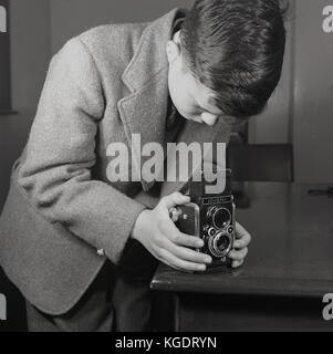 1950er Jahre, historische, England, UK, eine elegant gekleidete Teenager trägt eine Jacke und Krawatte verwendet eine Tabelle Ecke für Stabilität, als er ein Bild mit einem ferrania Elioflex Kamera nimmt. Dies war ein italienischer Film Kamera, die das Aussehen eines TLR (Twins Lens Reflex) Rolleiflex, die professionelle Kameras ihrer Tag, aber dies war nicht eine als Sucher war kein echter Reflex finder imitiert. Es wurde daher als "pseudo-TLR', war aber in dieser Zeit bekannt. Stockfoto