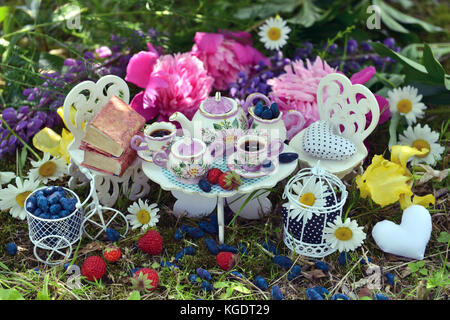 Teeservice, kleine Möbel, Beeren und Blumen auf dem Gras. Verrücktes Teeparty-Konzept. Wunderschöne Grußkarte Stockfoto