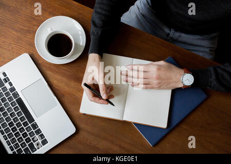 Styled lifestyle Foto von einem Mann in einem dunklen, hölzernen Schreibtisch zu Hause oder im Cafe schreiben in einem Notebook mit einem MacBook Laptop mit einer Kaffeetasse und Untertasse. Stockfoto