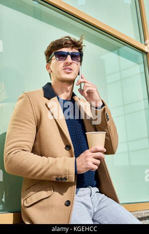 Portrait von gutaussehenden jungen Mann mit Sonnenbrille sprechen telefonisch bei Kaffee Pause draußen im Sonnenlicht Stockfoto