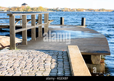 Holz- Coastal walkway einen heraus in das Meerwasser biegen. Herbst Landschaft im Hintergrund. Stockfoto