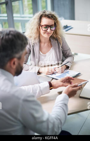 Attraktive mittleren Alters white collar worker in Brillen am Schreibtisch in der modernen Großraumbüro sitzen und die Analyse von Finanzdaten mit bärtigen Colle Stockfoto