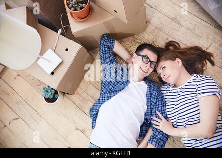 Reizende junge Paar liegend auf hölzernen Boden Ihrer neuen Wohnung und den Austausch von Ideen über die Gestaltung des Innenraums Wohnzimmer, Stapel von Kisten st Stockfoto