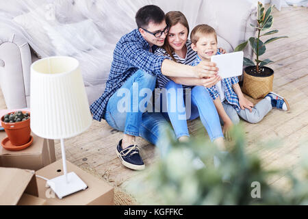 Adorable Familie von drei zusammen im Wohnzimmer der Wohnung und unter selfie auf digitalen Tablet versammelt, Innenarchitektur und beweglichen Kästen Stockfoto