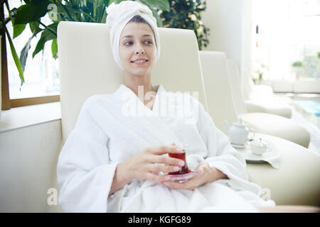 Porträt der schönen jungen Frau lächelnd entspannen in der Lounge Chair von Schwimmbad, Bademantel und Handtuch auf dem Kopf Stockfoto