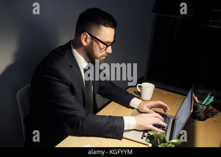 Aufmerksame bärtigen Mann in Gläsern, die am Projekt spät in der Nacht ist das Verfassen von Nachrichten, sich windende e-mails mit Laptop. Vergessen kalte Tasse Kaffee Stockfoto