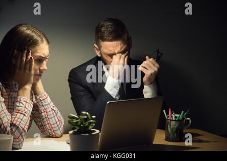 Zwei Kollegen treffen in dunklen Abend im Büro, um ein Problem zu lösen. Frist nähern. Mann nahm seine Brille ab und rieb die Augen, Frau sieht in lapt Stockfoto