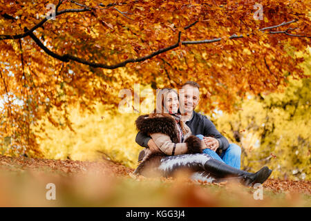 Schöne lächelndes Paar beim im sonnigen Wald im Herbst Farben. Sie auf der falls Blätter sitzen und auf Kamera. Stockfoto