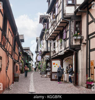 Rue des Remparts, eine gepflasterte, mittelalterliche Straße in Riquewihr im Elsass. Einheimische gehen über ihr tägliches Leben zwischen den bemalten, Fachwerkhäusern und Stockfoto