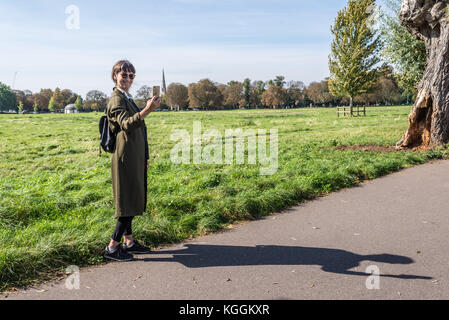 Junge Frau mit langen dunklen Olivgrün hellen Mantel bekleidet, kleine Leder Rucksack und schwarze Turnschuhe tragen Sonnenbrillen lächelnd und selfie in Stockfoto