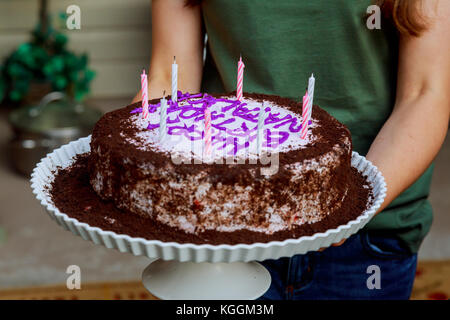 Junge Frau hält Teller mit leckeren Geburtstagskuchen gegen defokussierten Lichter Happy Birthday Kerzen auf Schokolade Kuchen Stockfoto
