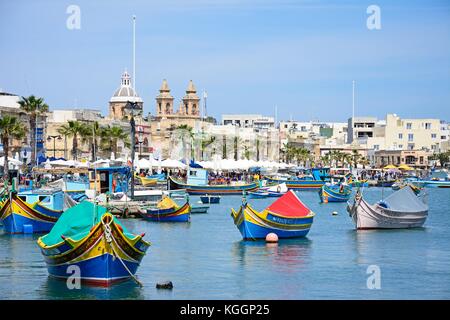 Traditionelle maltesische Dghajsa Fischerboote im Hafen mit der Pfarrkirche der Madonna von Pompei nach hinten, Marsaxlokk, Malta, Europa. Stockfoto