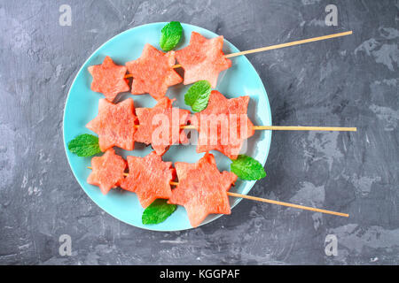 Wassermelone in Form von Sternen auf Spieße mit Blättern der Minze liegt auf einer Platte. Die blaue Teller ist wie eine Rakete im Weltraum. top View Stockfoto