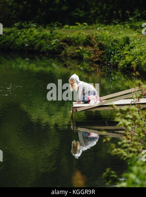 Süße kleine Mädchen sitzen auf hölzernen Pier, mit Blick auf den Bergsee. Stockfoto