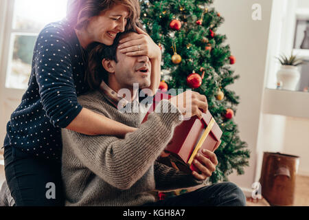 Ein paar glückliche Momente verbringen während Weihnachten zu Hause feiern. Lächelnden jungen Frau die Augen ihres Partners während über Weihnachten gif Übergabe Stockfoto