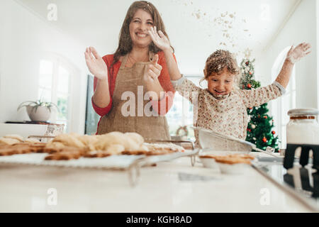 Glückliche Mutter und Tochter splash Mehl in die Luft, während Sie Cookies. Gebackene Kekse, Muffins und Lebkuchen am Küchentisch mit verzierten Christm Stockfoto