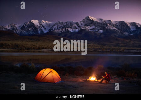 Camping bei Nacht. Männliche Touristen sitzen in dem beleuchteten Zelt in der Nähe des Lagerfeuers unter dem atemberaubenden Abendhimmel bei Sonnenuntergang in einer Berggegend. Schneeberg im B Stockfoto