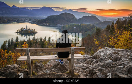 Bled, Slowenien - Runner Woman erholt sich und genießt die schöne Herbstansicht und den farbenfrohen Sonnenaufgang von Lake Bled auf einer auf einem Hügel sitzenden Bank Stockfoto