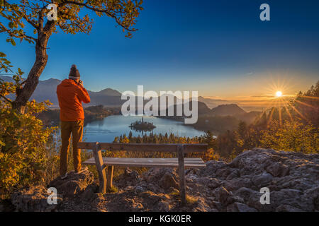 Bled, Slowenien - Fotograf Reisende, der orangefarbene Jacke und Hut trägt, fotografiert den Panoramablick auf den Herbstaufgang der Julischen Alpen, den Bleder See mit Stockfoto