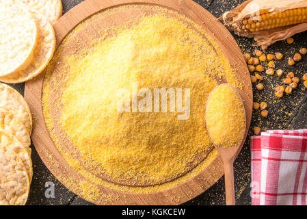 High Angle View Bild mit einem Stapel von Mais Mehl auf eine runde Holzplatte mit Maiskolben, Körner und Gepuffter mais Kuchen umgeben, auf einem schwarzen Holztisch Stockfoto