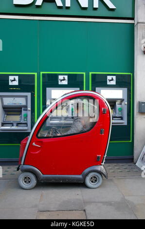 Mann in Rot Shoprider Mobilität scooter mit Geldautomaten Geldautomaten außerhalb Lloyds Bank in London, England Stockfoto