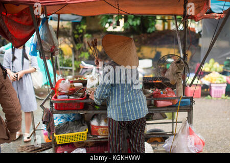 - Selektiver Fokus - nicht identifizierte Frau mit typischen vietnamesischen konischen Hüten verkaufen frisches Gemüse und Fleisch Spieße auf einem Straßenmarkt in Hoi An, Vietnam Stockfoto