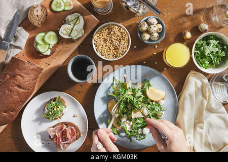 Zugeschnittenes Bild des Menschen mischen Salat mit Gabel und Messer in die Platte auf dem Tisch Stockfoto