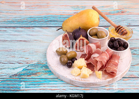 Käseplatte mit Scheiben Schinken, Käse, Oliven, Birnen, Honig und Feigen auf weißem Holz Schneidebrett aus Holz auf blauen Hintergrund. C Stockfoto