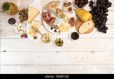 Die Gemeinkosten der Käseplatte mit verschimmelten Käse, Schinken, eingelegten Pflaumen, Oliven, Trauben, Feigen, Honig, Birne, Muttern auf weißem Holz- Hintergrund. Top Stockfoto