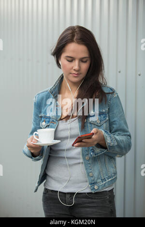 Schöne Frau mit Mobiltelefon, während Sie Kaffee Tasse gegen metallische Wand Stockfoto