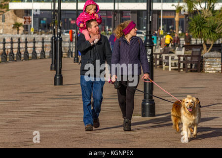 Eine Familie mit ein Paar, mit Ihrem Kind auf den Schultern des Mannes, und ihr Hund, zu Fuß entlang der Promenade von Torquay auf einem kühlen, aber sonnigen November Tag. Torquay, Devon, Großbritannien. November 2017. Stockfoto