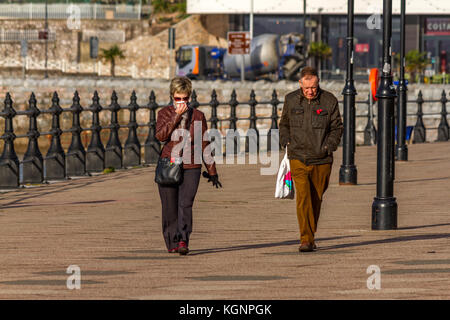 Ein älteres Ehepaar zu Fuß entlang der Promenade, einem kühlen, aber sonnigen Tag bringt Menschen in Torquay, November 2017. Stockfoto