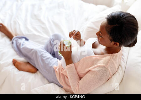 Schwangerschaft, Menschen und Rest Konzept - glücklich schwanger african american woman drinking grünes Gemüse Saft oder Smoothie in Bett zu Hause Stockfoto