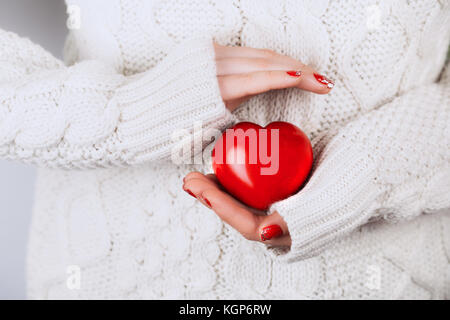 Die frau Hände mit schönen genaue Maniküre sorgfältig aufbewahren Rotes Herz. Weiße gemütlich gestrickten Pullover als Hintergrund. Konzept zu geben, die ihnen helfen, die Stockfoto