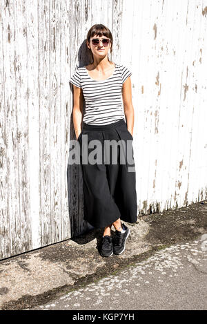 Hipster Frau mit Sonnenbrille in städtischen Minimal Style gekleidet mit Schwarz gestreiftes T-Shirt, Schwarz Hose mit weitem Bein und Turnschuhe. Schuß auf eine Ost Stockfoto