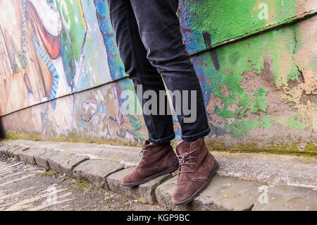 Modell trägt enge schwarze Jeans Skinny Hose und Brown suede Ankle Boots und Wandern vor einer Graffiti-wand. Konzentrieren Sie sich auf die Beine und Schuhe. Oberfläche Stockfoto