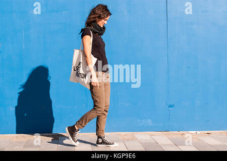 Junge Mädchen in casual braun Chinos, schwarze Turnschuhe und T-Shirt zu Fuß auf einer Straße mit blauen Wand im Hintergrund gekleidet. Stockfoto