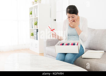 Junge attraktive Frau hat Paket und Sitzen auf einem Sofa öffnen Paket finden Kleidung vorhanden Gefühl auf Geburtstag überrascht. Stockfoto